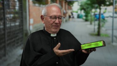 Siyah cüppeli kıdemli rahip elinde İncil ile sokak ortasında neşeli bir ifadeyle ve gündüz vakti şehir ortamında davetkar bir jest yapıyor.