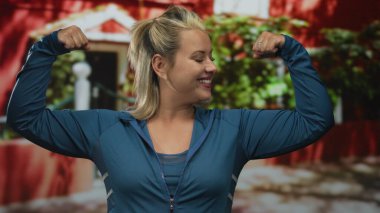 Sarışın kadın iki koluyla kaslarını esnetiyor. Evin girişinde mavi spor ceket giyiyor. Kendine güveni tam..