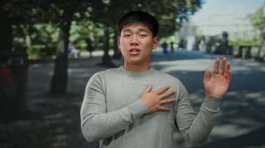 Gri kazaklı genç Çinli adam, yeşil ağaçlar ve gölgelerle çevrili güneşli bir parkta el hareketleriyle duygularını ifade ediyor..