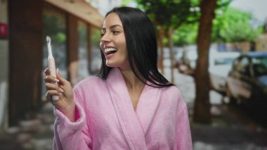 Pembe bornozlu gülümseyen kadın elektrikli ve manuel diş fırçalarını arka planda yeşil ağaçlar ve bulanık arabalarla şehir caddesinde tutuyor..