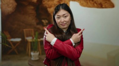 Kapalı bir yerde kırmızı ceketli çapraz kollarıyla poz veren bir kadın, rahat bir otel arka planına karşı kurulmuş, yön duygusunu ve karar verme mekanizmasını aktarıyor..