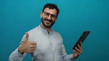 Sakallı İspanyol adam mavi arka planda tablet kullanıyor. Başparmak işareti yapıyor ve gülümsüyor.