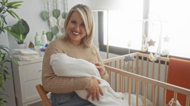 Sarışın kadın, güneşli bir yatak odasında elleri beyaz bir battaniyeye sarılı yeni doğmuş bebeği nazikçe kucaklar..