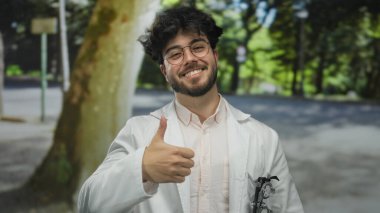 Sakallı, gözlüklü ve beyaz önlüklü İspanyol bir adam yeşil bir açık hava parkında baş parmağını kaldırıyor..