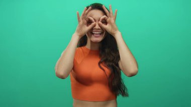 Portakal rengi bluzlu genç İspanyol kadın stüdyoda dürbün işareti yapıyor..