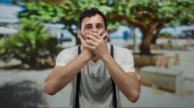 Sakallı İspanyol bir adam, arka planda yeşillik olan bir sokakta ağzını kapalı tutarak, canlı bir şehir ortamında çeşitli duygular sergileyerek şaşkınlığını dile getiriyor..