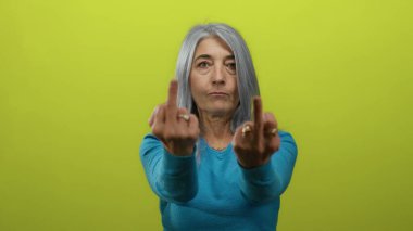 Yaşlı gri saçlı kadın müstehcen bir jest yapıyor. İki orta parmağı da havada, parlak sarı arka plan duvarına karşı duruyor. Sert bir ifadeyle..