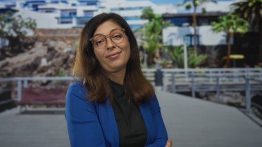 Gözlüklü ve kolları çaprazlanmış mavi ceketli bir kadın sokak geçidinde başını eğiyor..