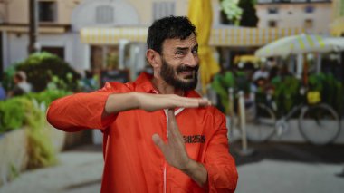 Turuncu hapishane tulumu giymiş İspanyol bir adam, dışarıda, sokakta hareketli bir jest yapıyor. Canlı bir arka plana karşı ciddi bir ifade takınıyor..