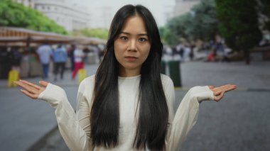 Kalabalık bir şehir pazarında, açık havada dikilen, günlük beyaz giysiler giyen, canlı bir caddede insanlar ve dükkanlar tarafından çevrili bir kadın..