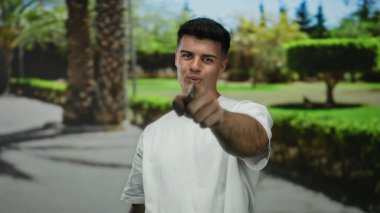 Genç adam, canlı yeşil ağaçlar ve güneş ışığının gölgelerinin olduğu açık hava parkına güveni ile işaret ederek doğal bir ortamda pozitif ve çekici bir tavır sergiliyor..