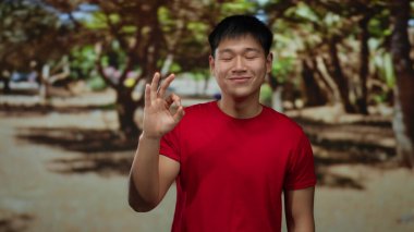 Kırmızı tişörtlü genç Çinli adam açık hava parkında iyi bir jest yapıyor..
