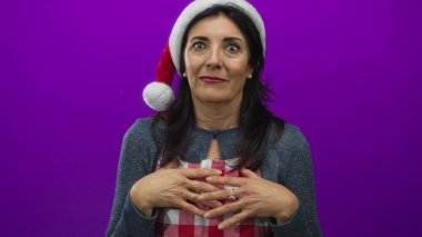 Orta yaşlı İspanyol kadın Noel Baba şapkası ve kırmızı önlük giyer ve mor stüdyoda avuçlarını açar..