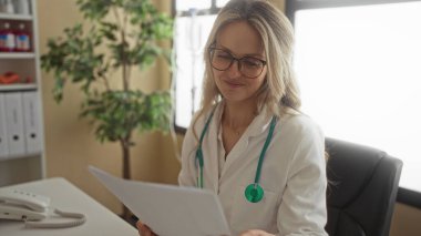 Steteskoplu kadın doktor klinikte çalışıyor. Kapalı yerlerde gözlük ve beyaz önlük takıyor. Tıbbi ekipman ve belgeler masanın üzerinde.