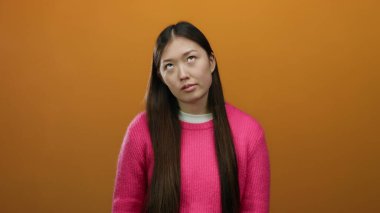 Pembe kazaklı, uzun saçlı bir kadın turuncu arka planda duruyor ve izole duvarın üzerinde derin derin bir ifade var..