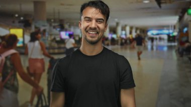 Genç İspanyol jest yapan bir adam elleri kapalı bir havaalanında gezginler ve bavullarla çevrili bir şekilde gülümsüyor..