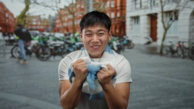 Genç adam heyecanlı bir şekilde mavi ve beyaz bir eşarp tutuyor. Şehrin hareketli caddesinde park etmiş bisikletlerle ve arka plandaki bulanık binalarla..