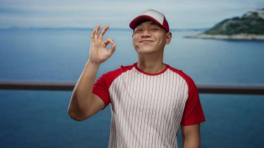 Beyzbol üniformalı genç adam deniz kenarındaki sahilde dikiliyor. Okyanus manzaralı hoş bir jest yapıyor.