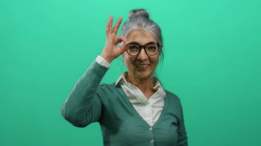 Gri saçlı yaşlı bir kadın, iyi bir jest yaparken özgüvenle gülümsüyor, canlı yeşil bir arka plan duvarına karşı duruyor, pozitifliği ve güveni temsil ediyor..