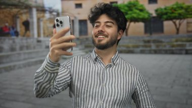 Genç adam, şık mimari ve yeşilliklerle çevrili, gösterişli İspanyol kültürü ve yaşam tarzını gösteren bir sokak ortamında akıllı telefonuyla selfie çekiyor..