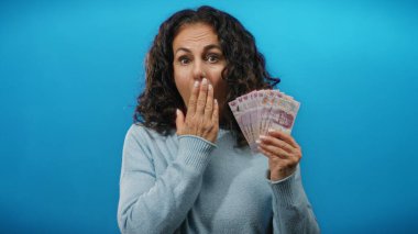 Orta yaşlı bir kadın, izole edilmiş mavi arka plana karşı sürpriz bir ifadeyle Meksika pezosu tutuyor..