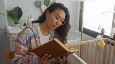 Gözlüklü genç İspanyol kadın, bir sandalyede oturur, kitap okur ve elinde çiçeklerin yanındaki bir bebek beşiği ve stüdyoda asılı bir cep telefonu vardır..