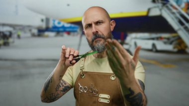 Havaalanı terminalinde makasıyla poz veren sakallı yakışıklı kel bir adam, açık havada stilist önlük giyerken açık uçuşu öneriyor..