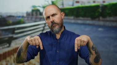 Sakallı, mavi gömlekli, şehir manzarası ortasında düşünceli bir yüz ifadesi yakalayan kel bir adam.