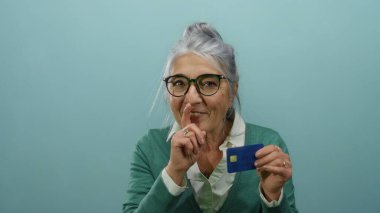 Mavi kredi kartı tutan, gözlüklü bir kadın mavi arka plana karşı sessiz bir jest yapıyor, kıdemli, düşünceli bir şekilde izole edilmiş, sağduyulu ya da mahremiyeti öneriyor..