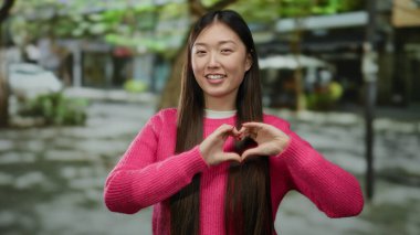 Pembe kazaklı bir kadın şehir sokaklarında elleri ile kalp şekli yapıyor. Neşenin, mutluluğun ve aşkın vücut bulmuş halidir..