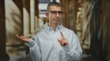 Beyaz adam el kol hareketi yapmamış ve avucunu açık havada uzatmış, gözlüklü ve çizgili gömlekli, bulanık bir arka plana karşı onay veya talimat iletiyor..