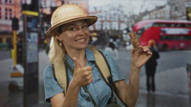 Atıştırmalık barını tutan kadın kaşif, kırmızı otobüs ve şehir binalarıyla dolu yoğun bir caddede başparmak kaldırıyor; mutluluk macerası keşfi.