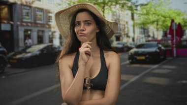 Kadın, saman şapka ve siyah bikinili kalabalık bir caddede ileri işaret ediyor; özgüven uyarısı.