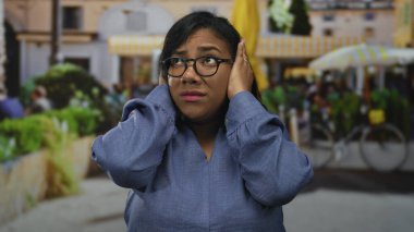 Gözlük takan Afrikalı Amerikalı kadın, gün boyunca hareketli şehir caddesinde kulaklarını elleriyle kapatıyor; rahatsız edici gürültü aşırı yüklenmesi.