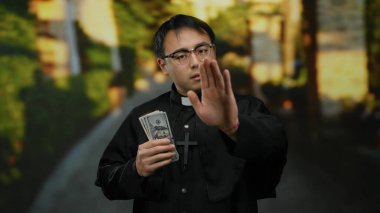 Asyalı rahip elinde para, gözlük ve siyah bornoz takıyor, el hareketleriyle inkar ediyor, dini ve finans konularını temsil ediyor..