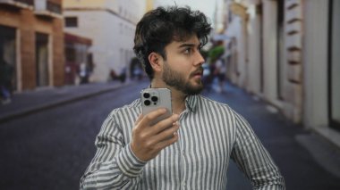 Sakallı genç adam şehir caddesinde akıllı telefon kullanıyor çizgili tişört giyiyor günlük ortamda çevreyi gözetliyor sokak deneyimlerini geliştiriyor..