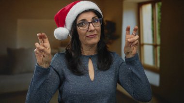 Oturma odasında Noel Baba şapkası takan bir kadın neşeli gülümsemeyle eğlenirken tırnak işareti yapıyor; bayram neşesi.