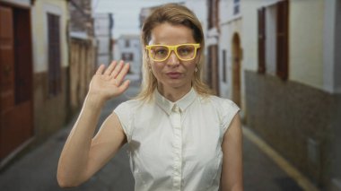 Beyaz gömlekli ve sarı gözlüklü bir kadın, taştan binalarla çerçevelenmiş güneşli eski kasaba sokaklarında el ele dolaşır..