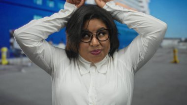 Genç İspanyol kadın deniz kıyısındaki bir limanda, büyük bir teknenin önünde elleriyle kulaklarını taklit ederek şakacı hareketler yapıyor..