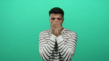 Genç adam elleri ile ağzını kapatıyor yeşil bir zemine karşı neşeli bir şekilde duruyor çizgili bir gömlek giyiyor duygularını ve şaşkınlığını gösteriyor..