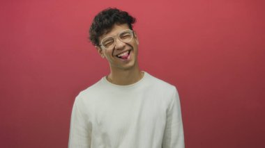 Yuvarlak gözlüklü ve beyaz gömlekli adam kırmızı stüdyoda dilini çıkarıp gözlerini kapatıyor..