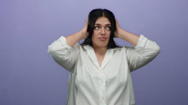 Elleri kulaklarının üzerinde olan bir kadın mor bir duvara dayanır, beyaz bir gömlek içinde hayal kırıklığını ifade eder; artı boyut genç bayan sade arka planda izole edilmiş, duygu ve beden pozitifliği iletir.