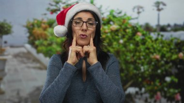 Noel Baba şapkası ve gözlük takan İspanyol orta yaşlı kadın sokakta şapkayı işaret ediyor; bayram neşesi.