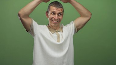 Çatı jesti yapan adam, elleri başının üzerinde, yeşil bir duvara yaslanmış, beyaz bir gömlek giymiş ve gülümsüyor..