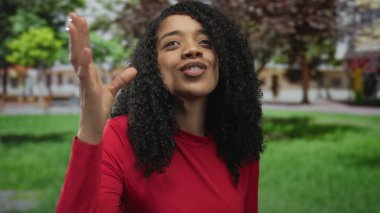 Kırmızı bluz giyen kadın dudaklarıyla öpüşüyor ve parkta yeşil çimlerin üzerine elini kaldırıyor..