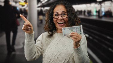 Uae istasyonundaki kapalı bir tren istasyonunda orta yaşlı bir kadın ve bulanık banliyö sakinleriyle çevrili, sürpriz bir ifadeyle bir Dirham banknotunu işaret ediyor..
