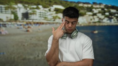 Sahilde bir plajda oturan genç adam kulaklık takıyor ve beyaz bir gömlek giyiyor. Düşünceli bir şekilde arka planda insanlar ve binalar var..