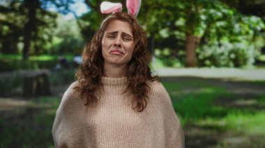 Parkta genç bir kadın üzgün bir ifadeyle tavşan kulağı takıyor, açık hava ortamında yemyeşil ile çevrili, doğada oyunculuk ve duygunun bir karışımını yakalıyor..