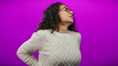 Kıvırcık saçlı İspanyol bir kadın canlı mor bir arka plan karşısında sırt ağrısı çekiyor. Sırtını izole bir ortamda tutarken rahatsızlık veriyor..