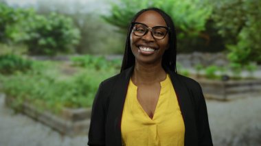 Açık hava parkında gülümseyen yeşil gözlüklü ve sarı gömlekli bir kadın mutluluğu ve kendine güveni yayıyor..